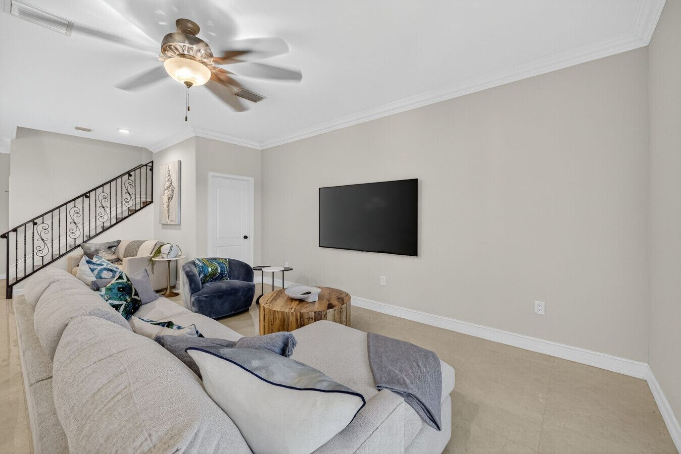 Un salon moderne avec un grand canapé, un fauteuil bleu, une table basse, un ventilateur de plafond, un escalier et un téléviseur à écran plat fixé au mur.