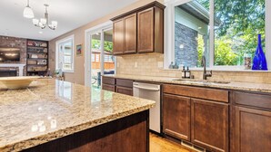 A modern kitchen with granite countertops, dark wooden cabinets, a built-in dishwasher, and a large window offering a view of the outdoors.