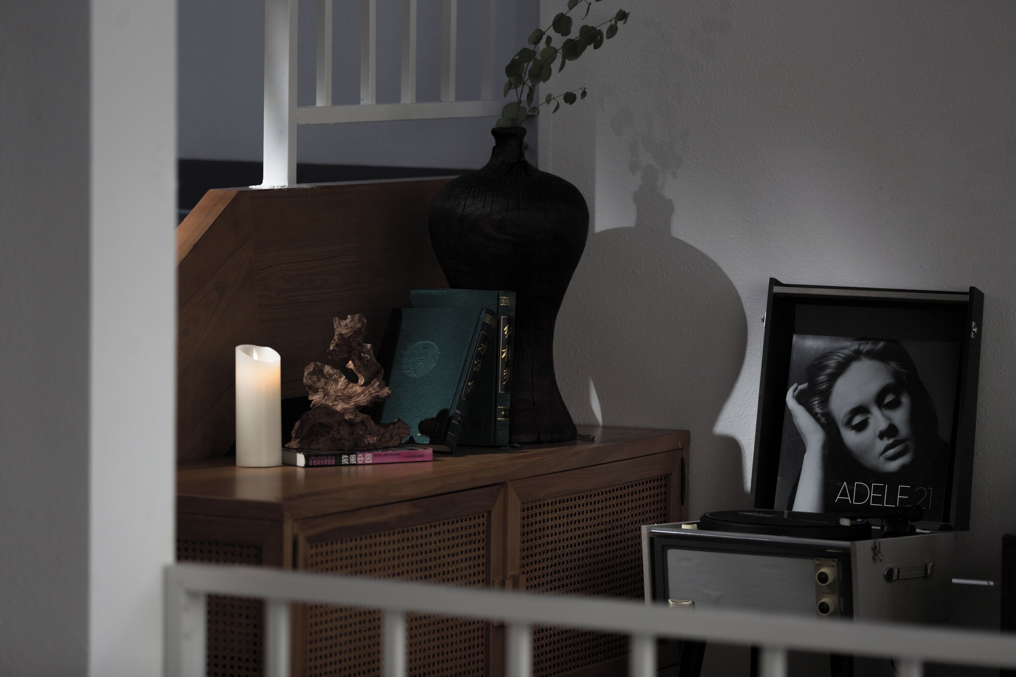 Una mesita auxiliar de madera con una vela blanca, un florero decorativo con una planta, un cuadro enmarco y algunos libros.