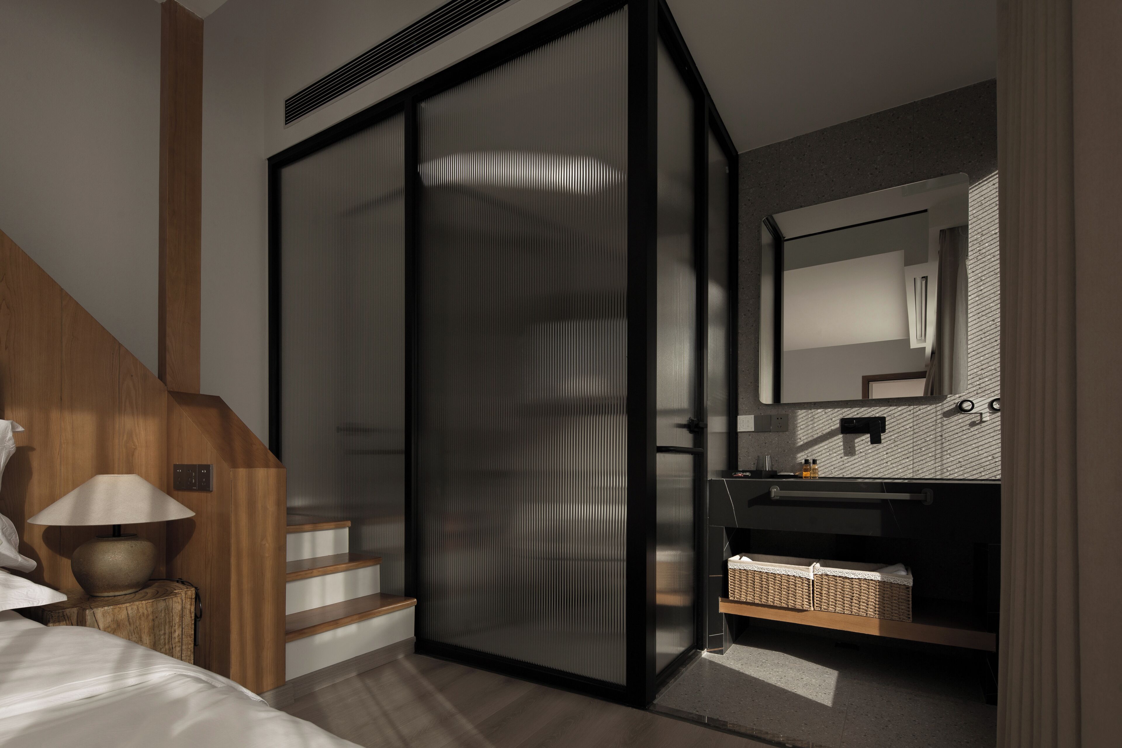 A modern bedroom with a wooden staircase, a glass partition, a bedside lamp, a nightstand, a bed, a bathroom with a mirror, sink, and a basket.