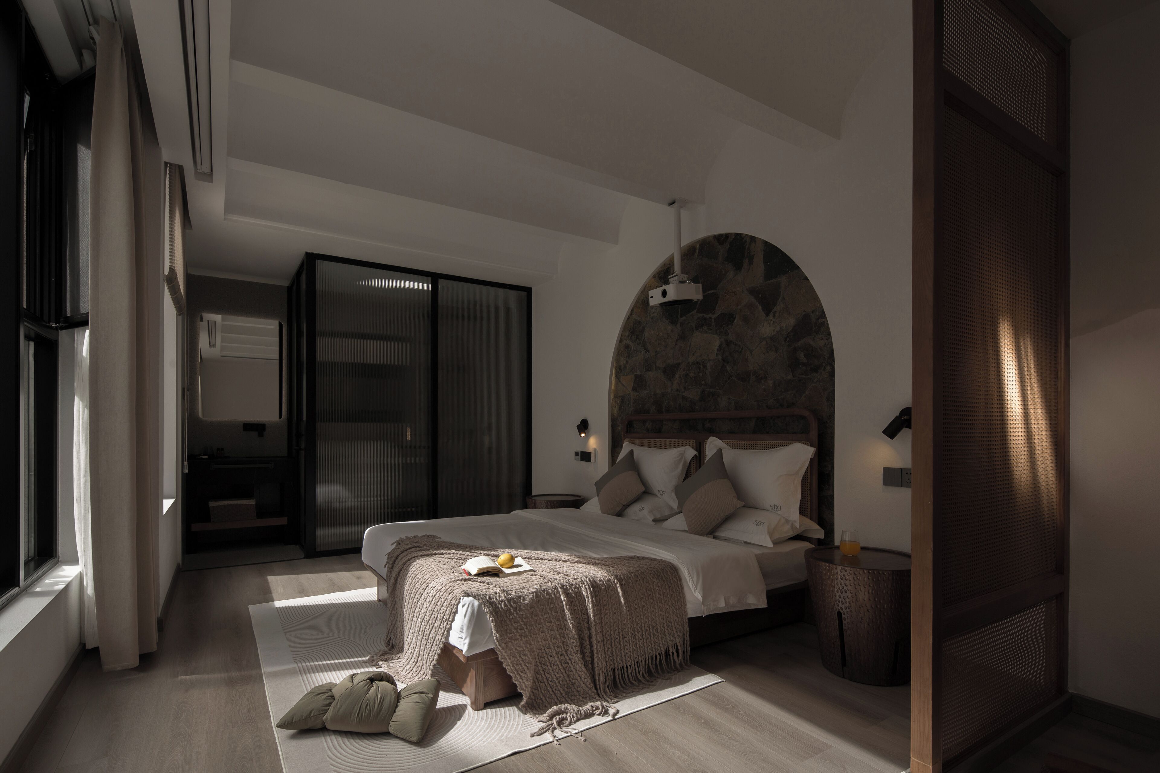 A modern bedroom with a large bed, a stone accent wall, a sliding glass door, and a small round table with a lamp.