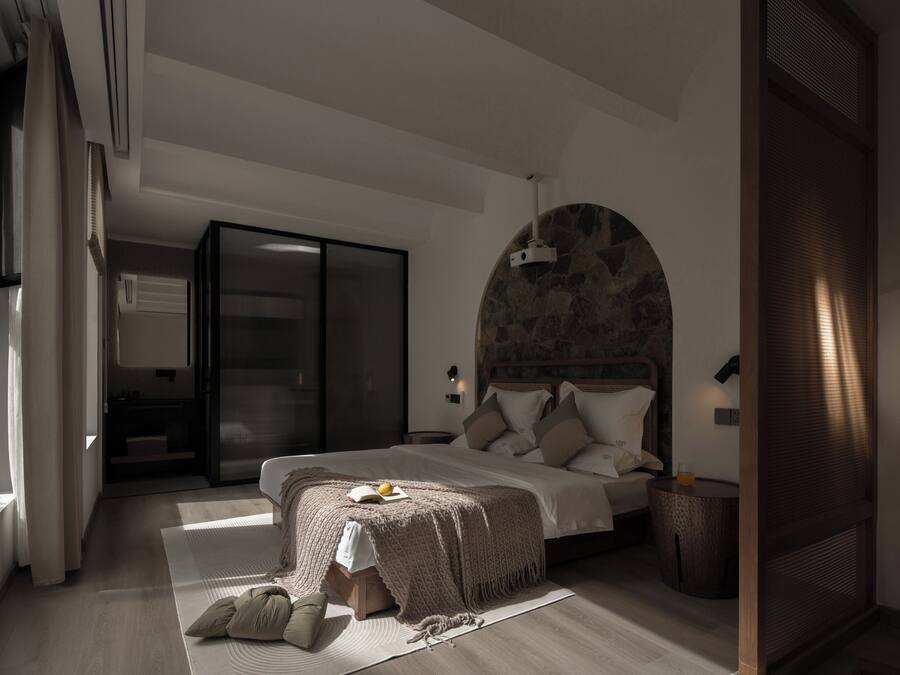 A modern bedroom with a large bed, a stone accent wall, a sliding glass door, and a small round table with a lamp.