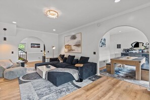A modern living room with a sectional sofa, a coffee table, and a dining table. There are two archways leading to other rooms. - OG Paradise | Private Pool & Hot Tub | Luxe Oasis (Wilmington)