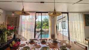 Un comedor con una mesa de madera dispuesta para una comida, ventanas amplias con cortinas y vistas a una piscina y follaje exterior.