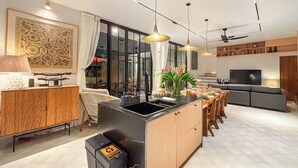 Una cocina moderna con una isla central, comedor con sillas, televisor y un amplio ventanal con cortinas.