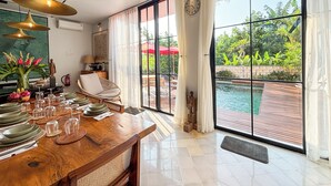Un comedor con una mesa de madera dispuesta para cuatro personas, una silla de mimbre y vistas a una piscina a través de amplios ventanales.