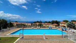 Ein rechteckiger Swimmingpool, umgeben von einem weißen Zaun, befindet sich in einer Wohngegend mit Häusern im Hintergrund und blauem Himmel.