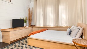 Une chambre à coucher avec un lit, un téléviseur posé sur une console en bois, une fenêtre avec des rideaux et une plante.