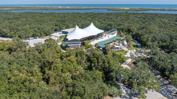 Vue aérienne d’un complexe hôtelier doté d’une structure blanche originale en forme de tente, entouré d’une forêt dense et avec une étendue d’eau en arrière-plan.
