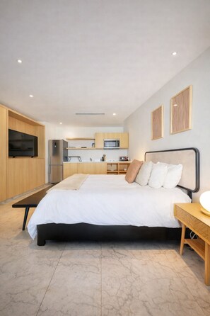 A modern bedroom with a large bed, bedside tables, a TV, and a kitchen area in the background. - Luxury Cabo Studio | Rooftop Pool, Hot Tub & Walk to Beach (Cabo San Lucas)