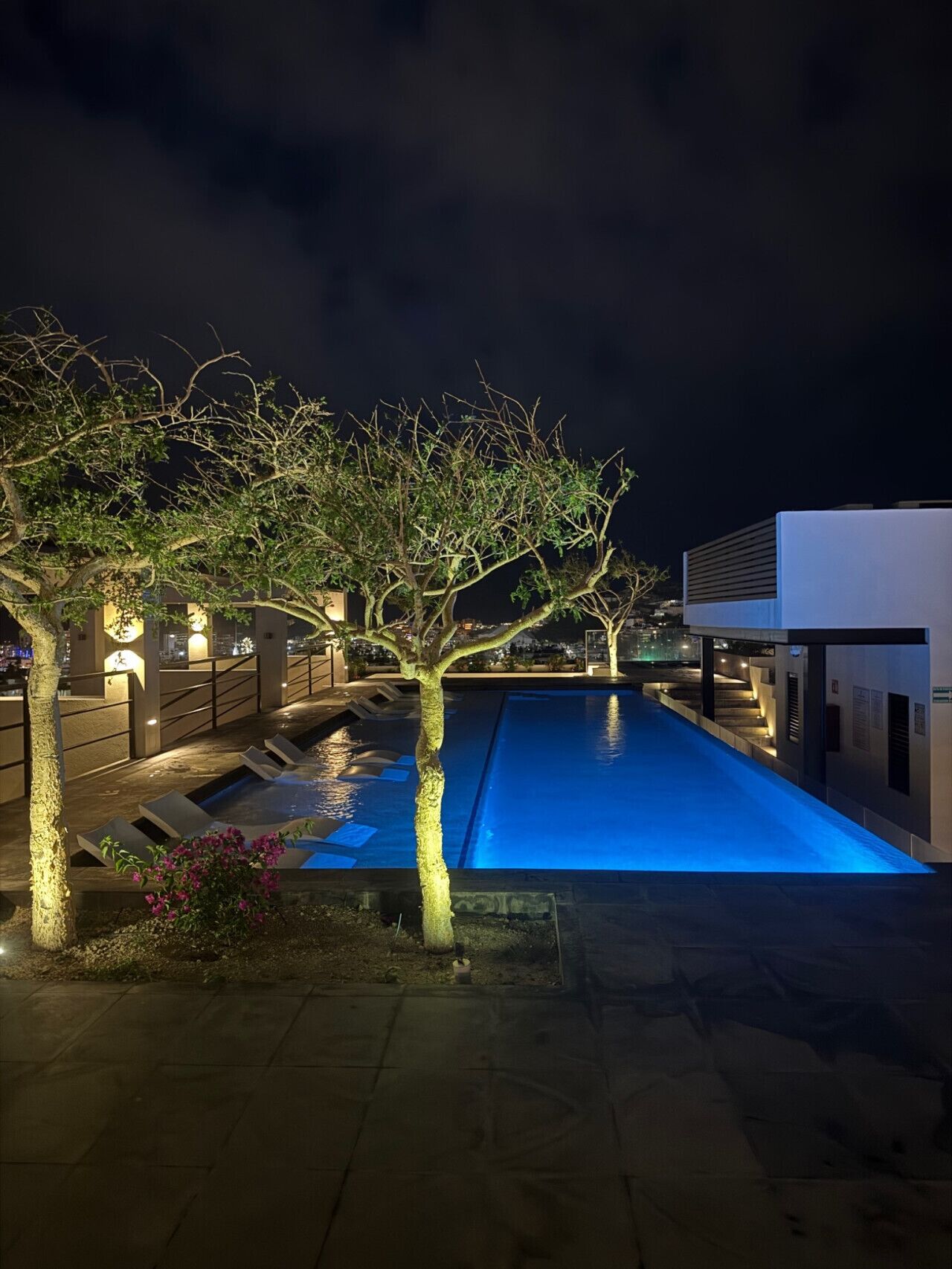Un área de piscina nocturna con diseño moderno, que incluye una piscina, sillas de descanso y una escalera que conduce a un nivel superior.