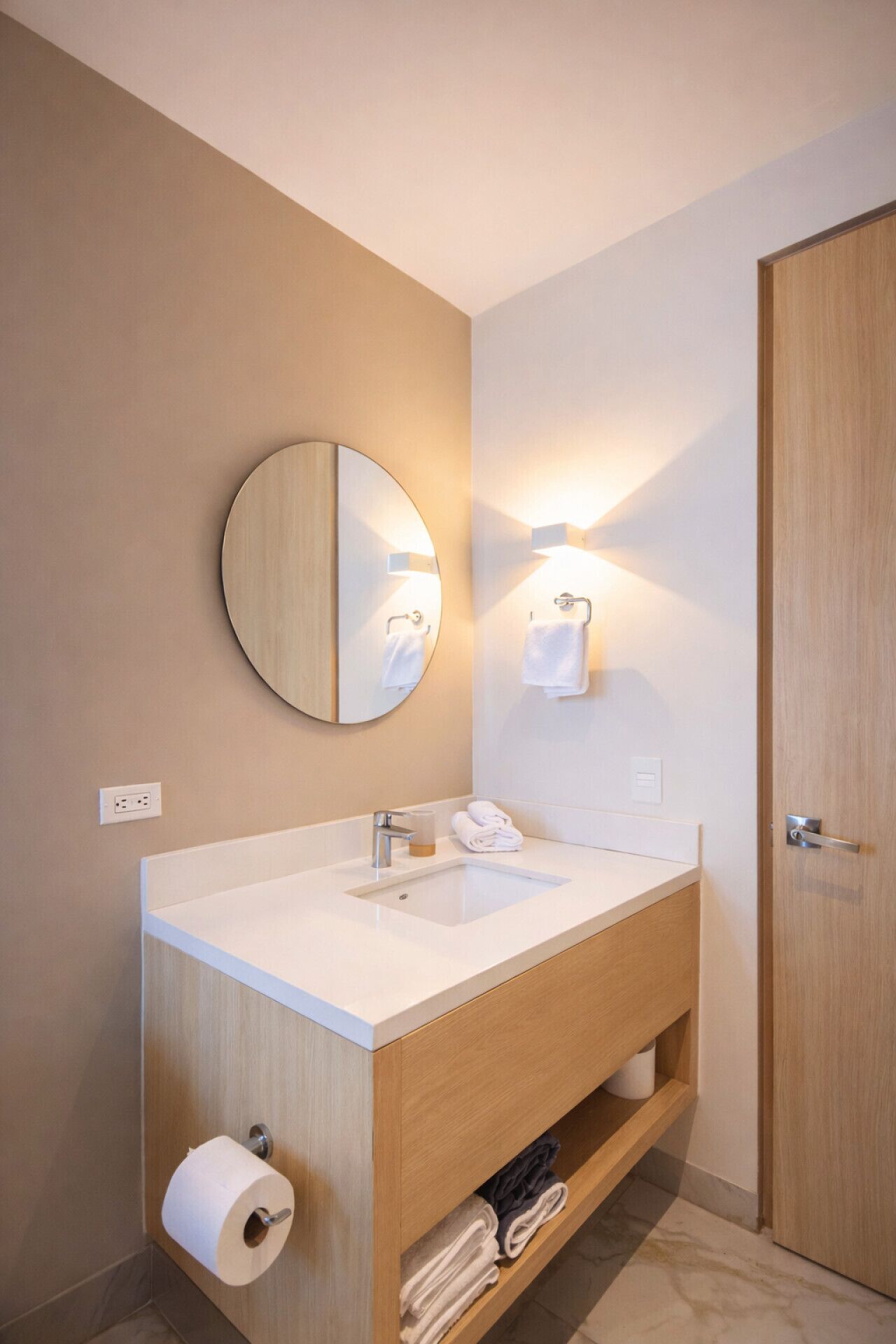 A modern bathroom with a wooden vanity, a round mirror, a wall-mounted light, and a door.