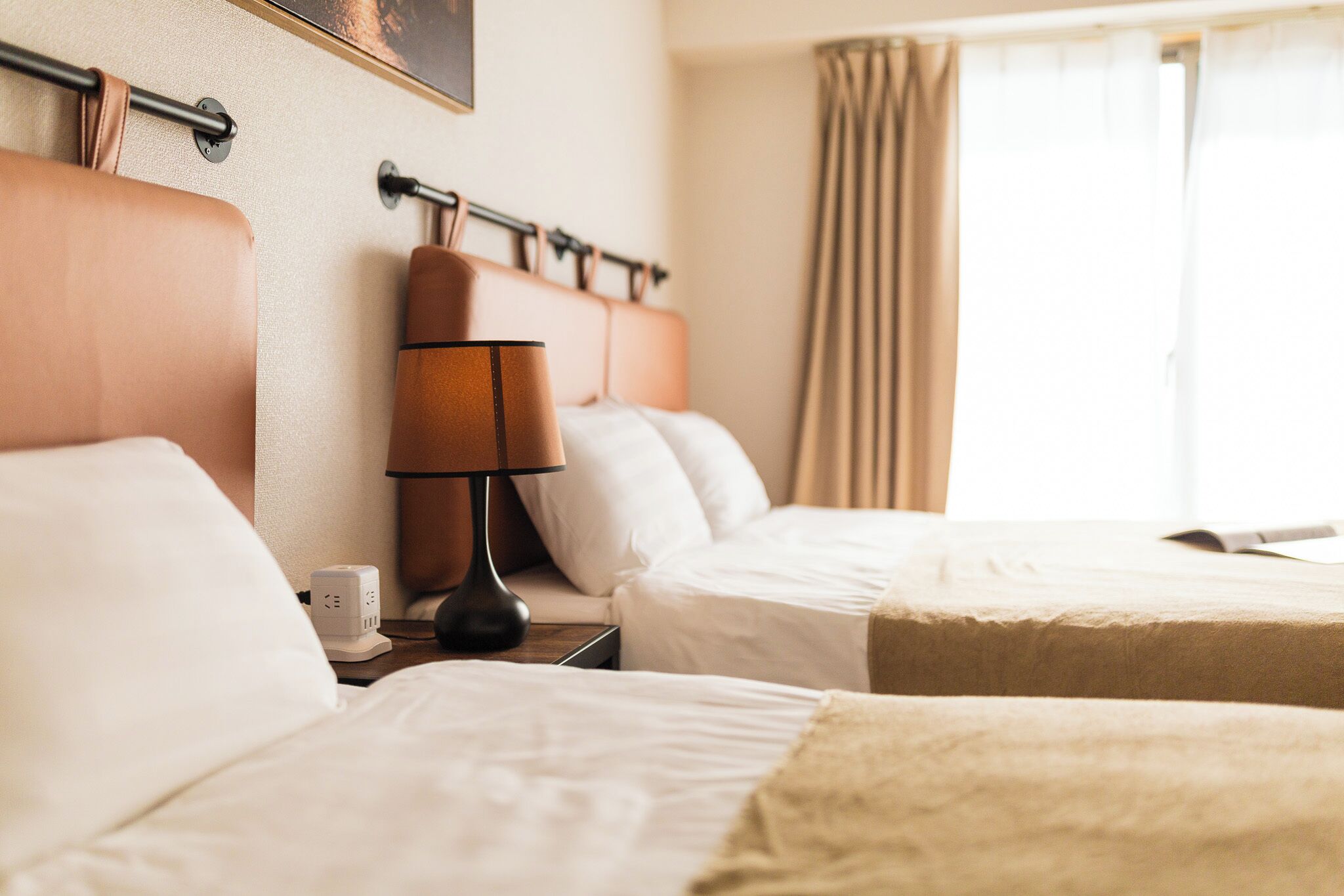 A hotel room with two beds, a nightstand with a lamp, a clock, and a window with curtains.
