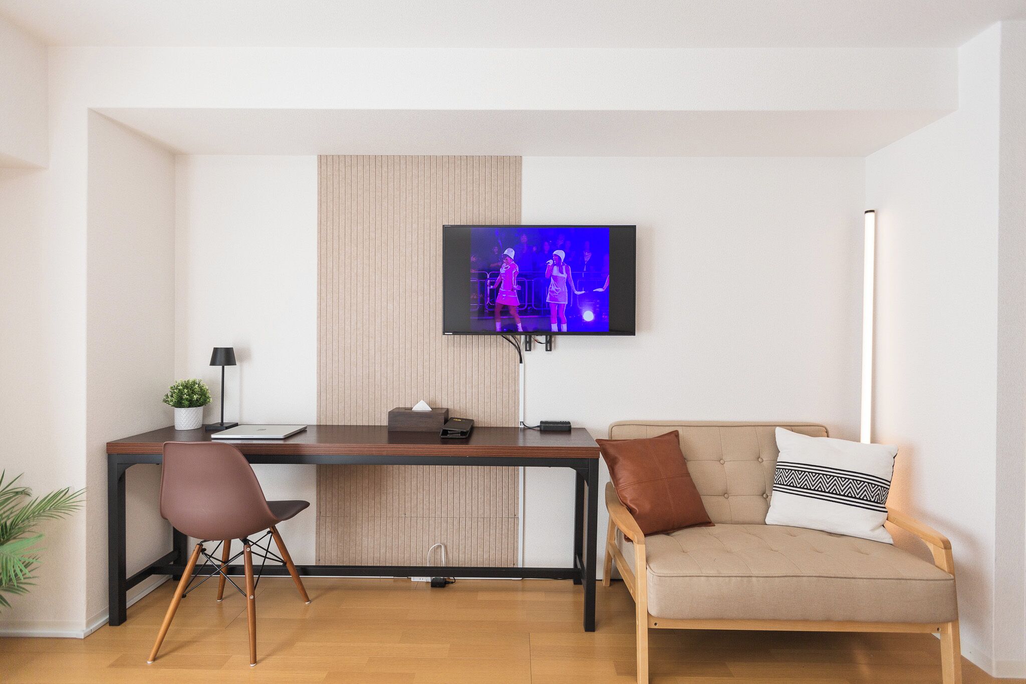 Un salón moderno con un televisor de pantalla plana montado en la pared, un escritorio de madera, una silla marrón, un sofá beige con cojines estampados y una pequeña planta en maceta.