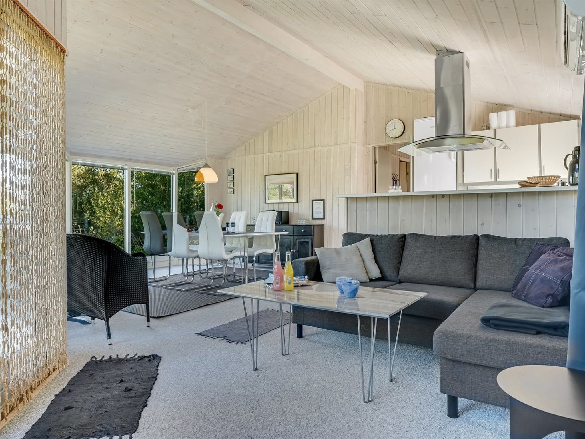 Ein modernes Wohnzimmer mit einer Couch, einem Couchtisch und Sesseln. Es gibt eine Küchenzeile mit einem Kühlschrank und einer Uhr an der Wand.