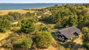 Une propriété située sur la côte, avec une grande maison dotée d’une terrasse et d’un espace salon extérieur, entourée d’une forêt dense et offrant une vue sur la mer.