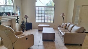 Ein Wohnzimmer mit Sofa, Sessel, Pouf, Kamin, Weihnachtstannenbaum und großem Fenster.