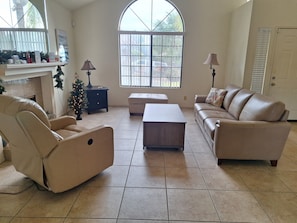 A living room with a sofa, armchair, and ottoman, a fireplace, a Christmas tree, and a large window. - Private Home in Greater Palm Springs, La Quinta 
Salt Water Pool and Hot Tub (La Quinta)