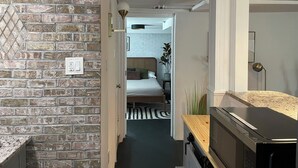 Un pasillo estrecho con una pared de ladrillo a un lado y una encimera de madera al otro. En el fondo, se ve una cama con cabecera y una mesita de noche con lámpara.