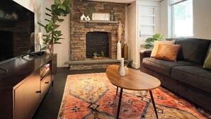 Una sala de estar con chimenea de piedra, un televisor de pantalla plana, un sofá oscuro y una alfombra estampada.