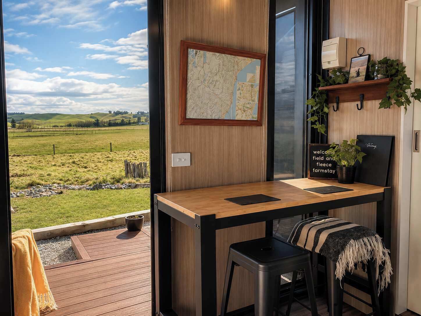 Una habitación con un escritorio de madera, dos taburetes negros, un mapa en la pared, un cartel de bienvenida y vistas a un campo verde.