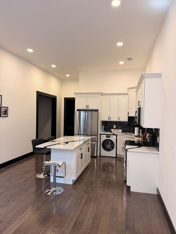 A modern kitchen with white cabinetry, a stainless steel refrigerator, a built-in washer and dryer, and a dining area with a white island and bar stools.