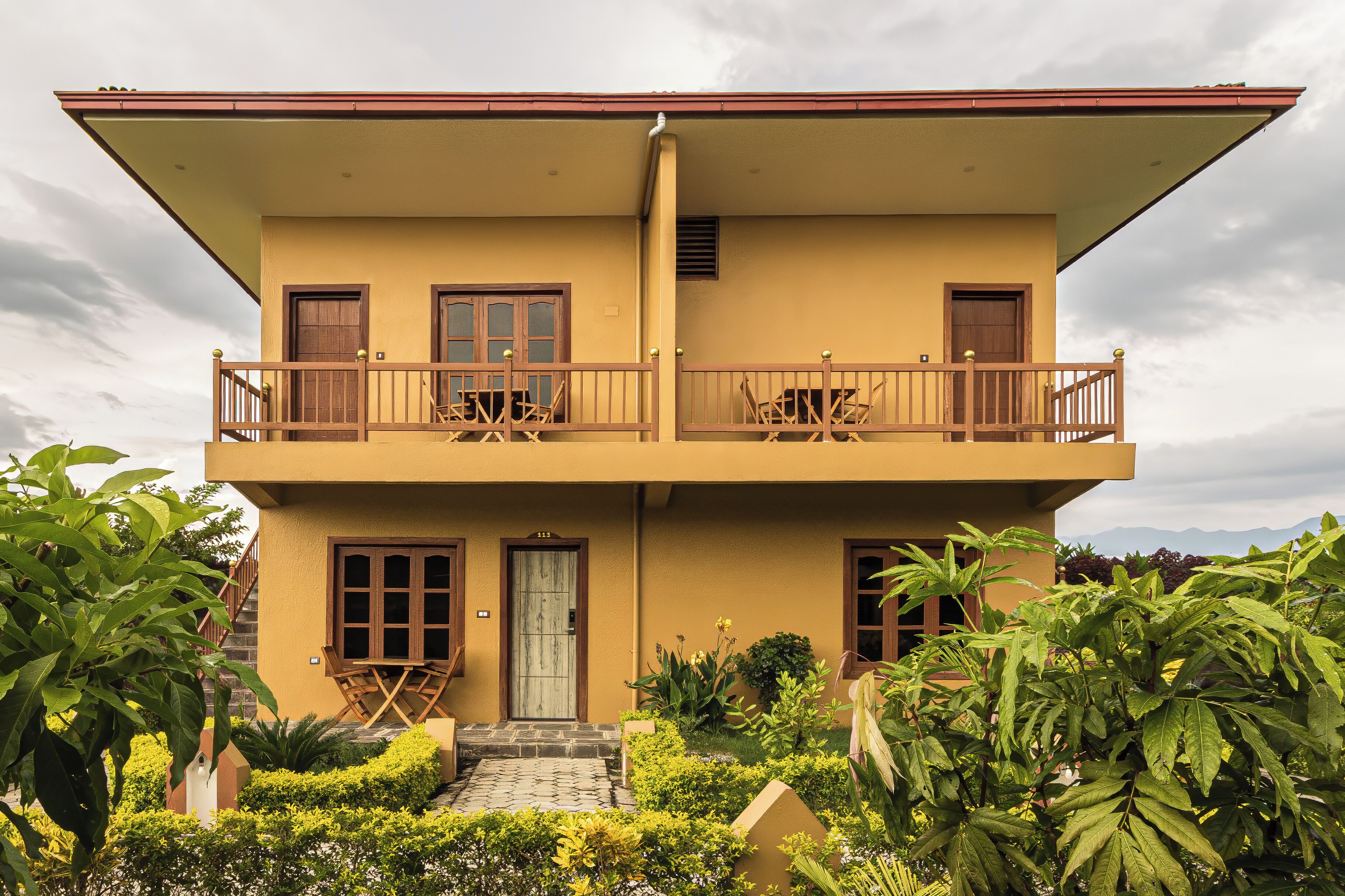 A two-story building with a balcony on the upper floor, a front door, and a small garden area with various plants.