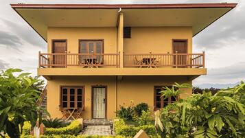 A two-story building with a balcony on the upper floor, a front door, and a small garden area with various plants.