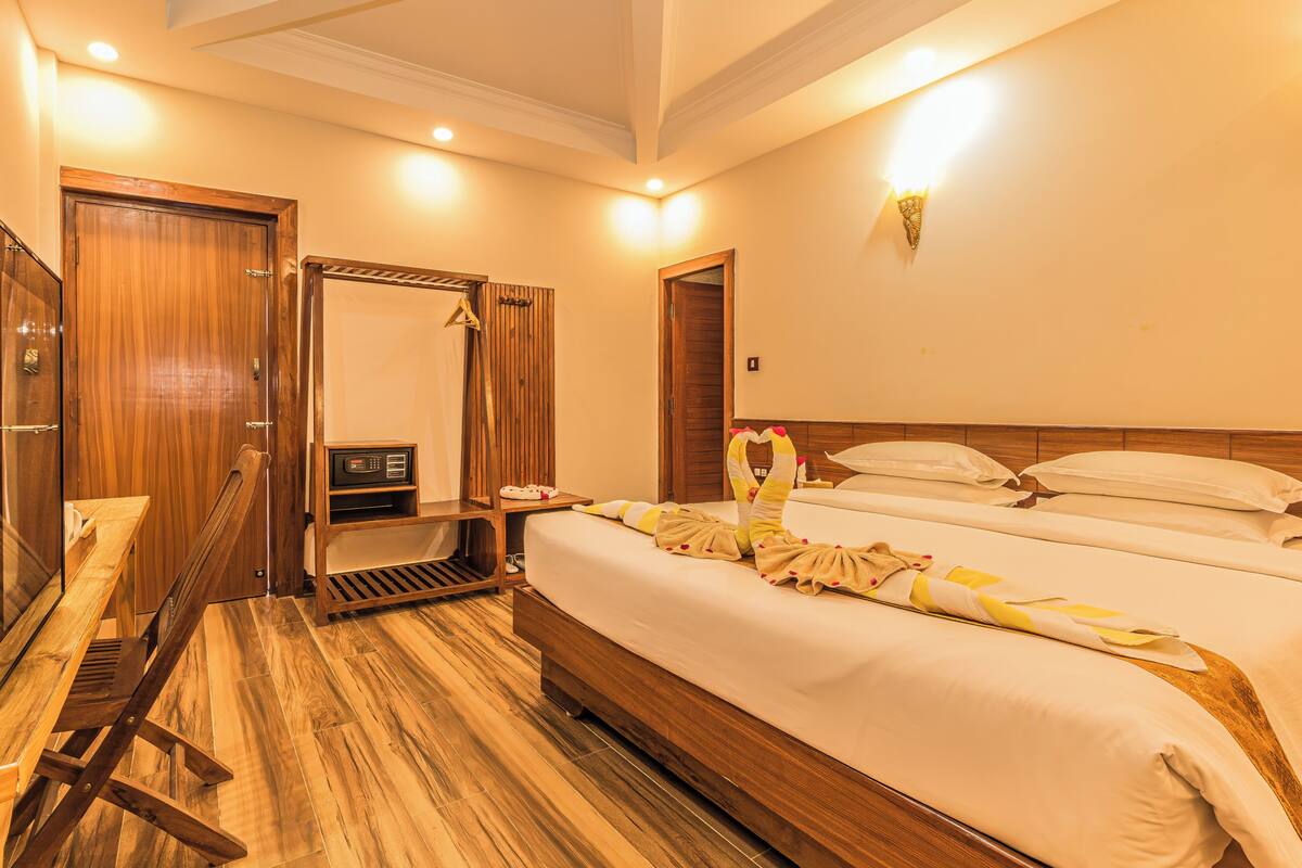 A hotel room with a wooden floor, a bed with white linens, a wooden wardrobe, a mirror, and a small table with a radio.