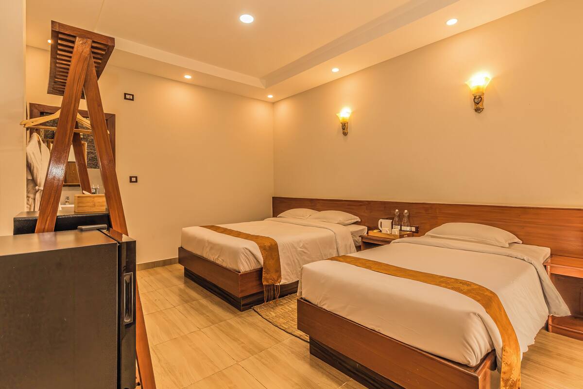 A hotel room with two beds, a wooden wardrobe, a desk, and a wall-mounted light fixture.