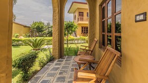 A covered outdoor area with a patio, two wooden chairs, a small table, and a view of a building with a balcony. - Munal and Mayur Resort (Kalika)