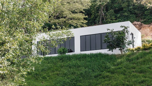 A modern, single-story building with a flat roof, large windows, and a minimalist design, surrounded by lush greenery and hills.