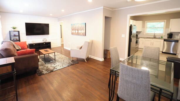 A modern living room with a sofa, coffee table, and a glass dining table with chairs. A flat-screen TV is mounted on the wall, and there is a painting above the sofa. - Cozy Decatur Bungalow with Beautiful Backyard (Decatur)