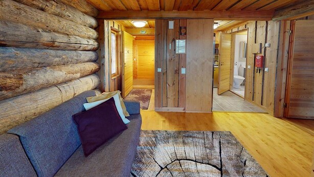 Interior de una cabaña de troncos con techo de madera, un sofá con cojines, suelo de madera y una puerta que da a otra habitación.