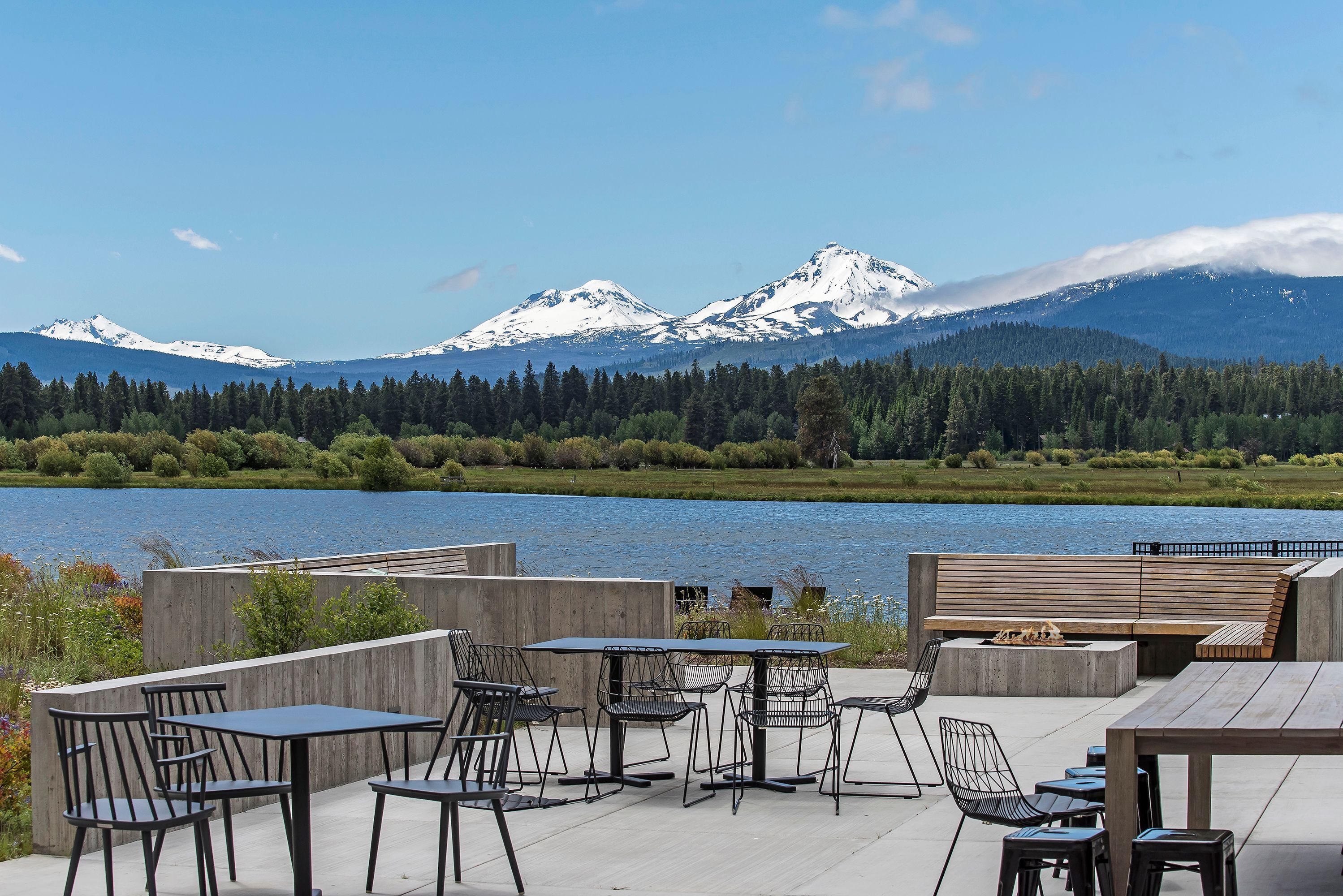 An outdoor seating area with tables and chairs, a fire pit, and a view of snow-capped mountains and a lake.