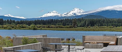 An outdoor seating area with tables and chairs, a fire pit, and a view of snow-capped mountains and a lake.