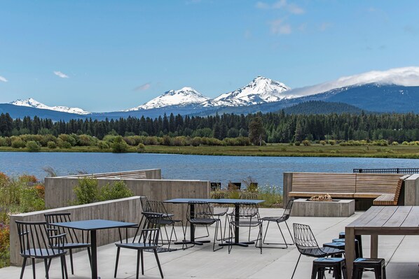 An outdoor seating area with tables and chairs, a fire pit, and a view of snow-capped mountains and a lake.