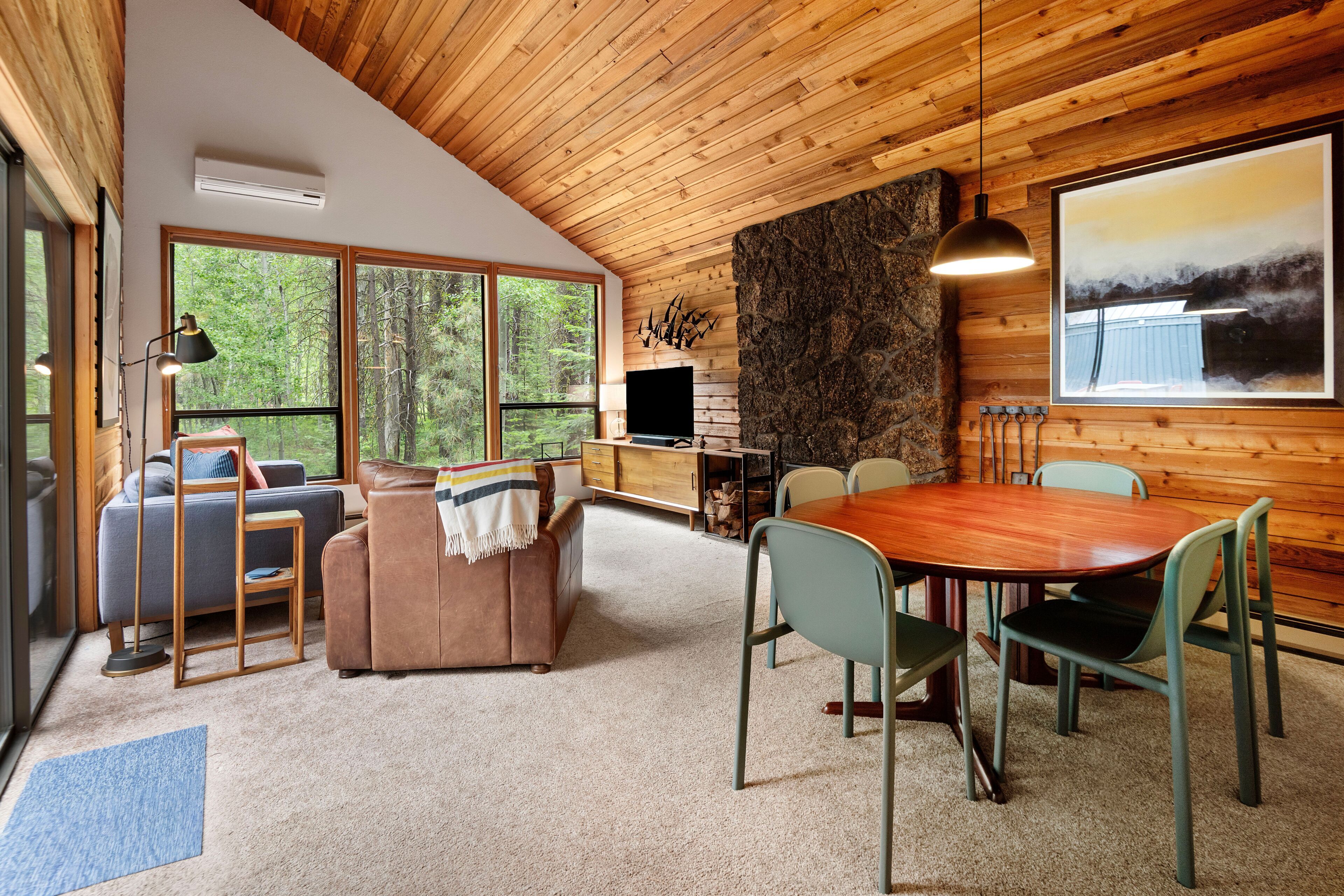 Un intérieur de chalet chaleureux avec un plafond en bois, de grandes fenêtres, une cheminée, un coin repas et un téléviseur.