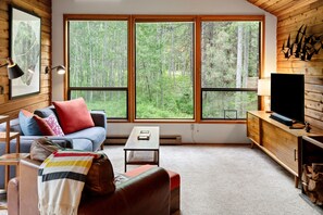 L’intérieur d’un chalet accueillant, avec une grande fenêtre, un canapé, une table basse, un téléviseur et une cheminée.