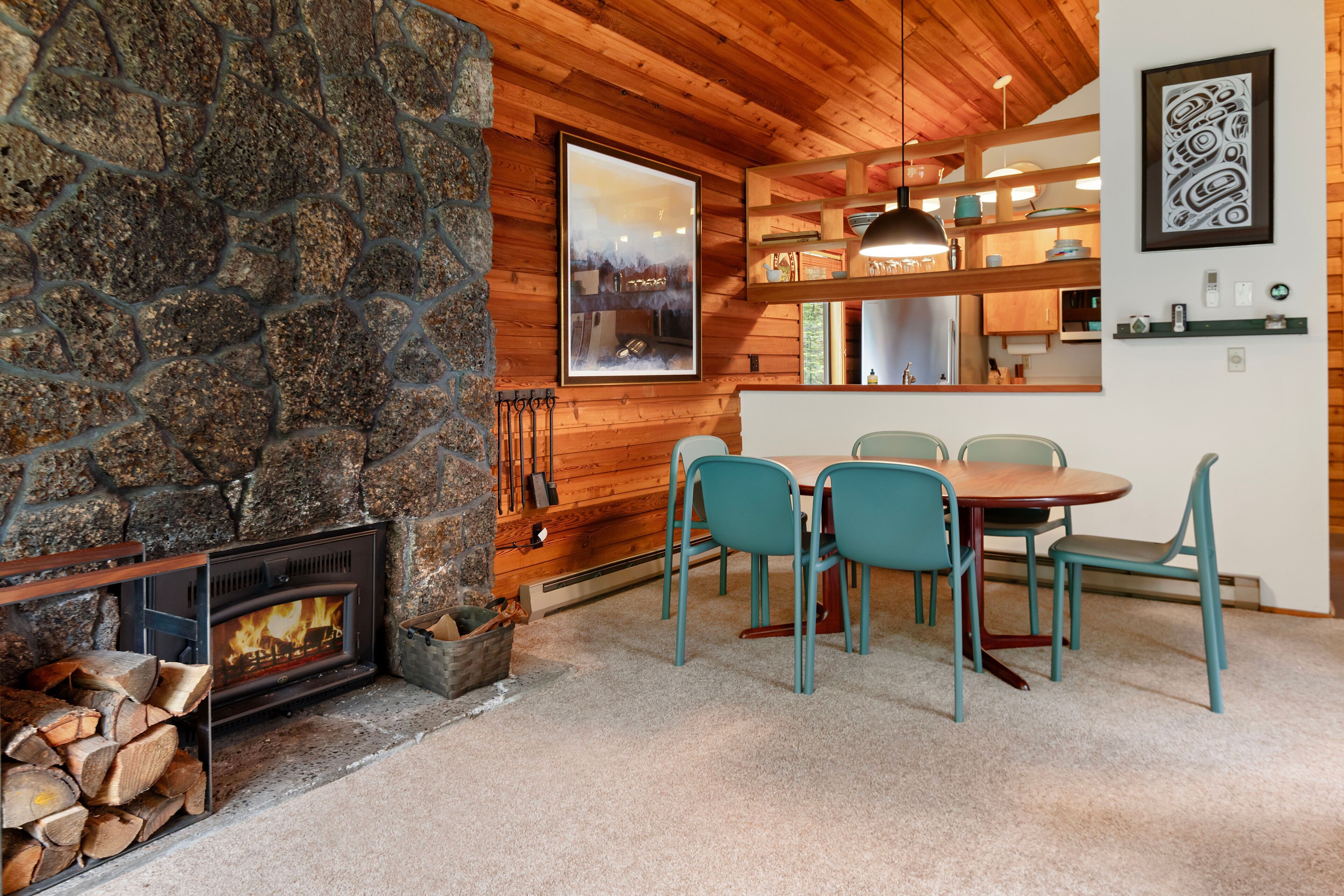 A stone fireplace, wooden ceiling, dining table with chairs, and a kitchen area with shelves and hanging lights.
