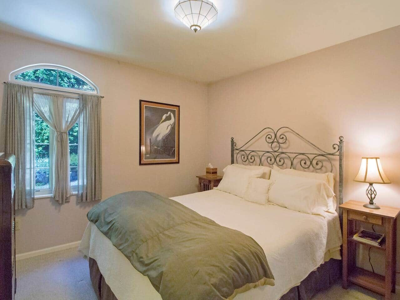 Ein Schlafzimmer mit Bett, Nachttisch mit Lampe, einem Fenster mit Vorhängen und einem gerahmten Bild an der Wand.