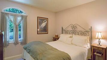 Ein Schlafzimmer mit Bett, Nachttisch mit Lampe, einem Fenster mit Vorhängen und einem gerahmten Bild an der Wand.