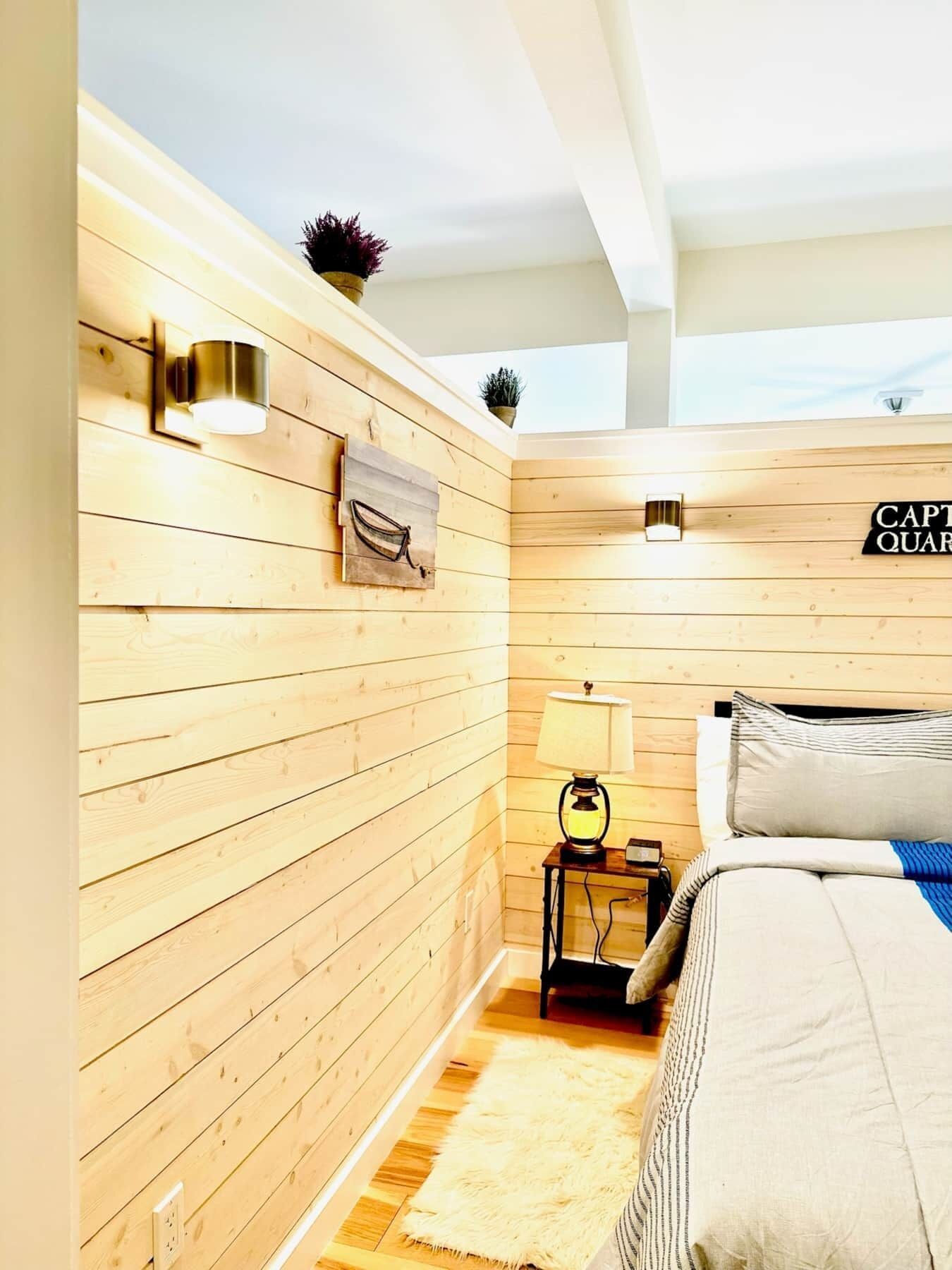 A room with wooden panel walls, a bed with a striped pillow, a nightstand with a lamp, and a wall-mounted light fixture.