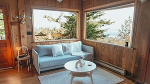 A cozy room with a light blue sofa, a small white table, and a view of trees and a body of water through large windows. - INSANE VIEWS! HOT TUB! (San Juan County)
