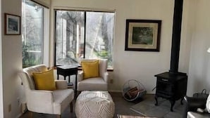 A living room with a sofa, armchairs, a table, a black stove, and large windows.
