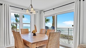 Un coin repas avec une table en bois et des chaises en osier, de grandes fenêtres donnant sur l’océan et un balcon agrémenté d’une balustrade.