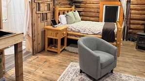A log cabin bedroom with a wooden bed, a small desk, a chair, and a large rug.