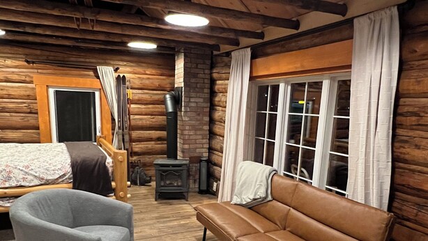 A log cabin interior with a bed, a gray armchair, a brown leather sofa, a patterned rug, and a wood-burning stove.