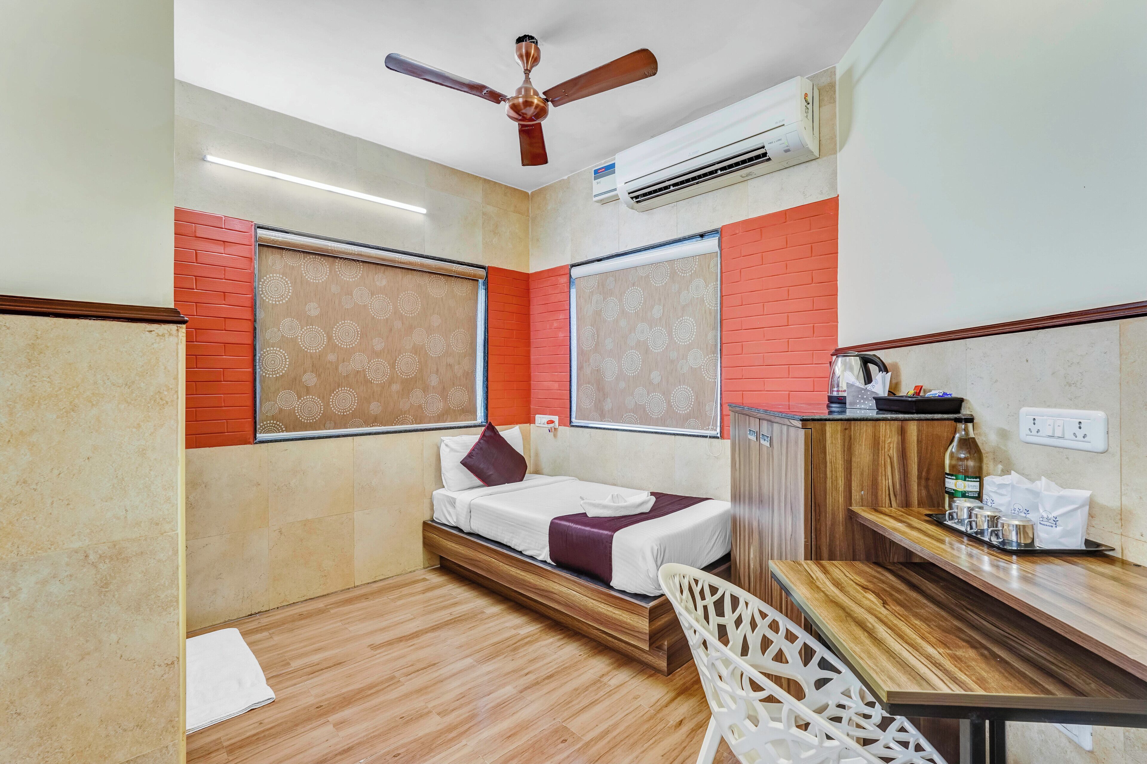 Habitación de hotel compacta con cama, escritorio y silla. Cuenta con ventilador de techo, unidad de aire acondicionado y ventana con vistas.