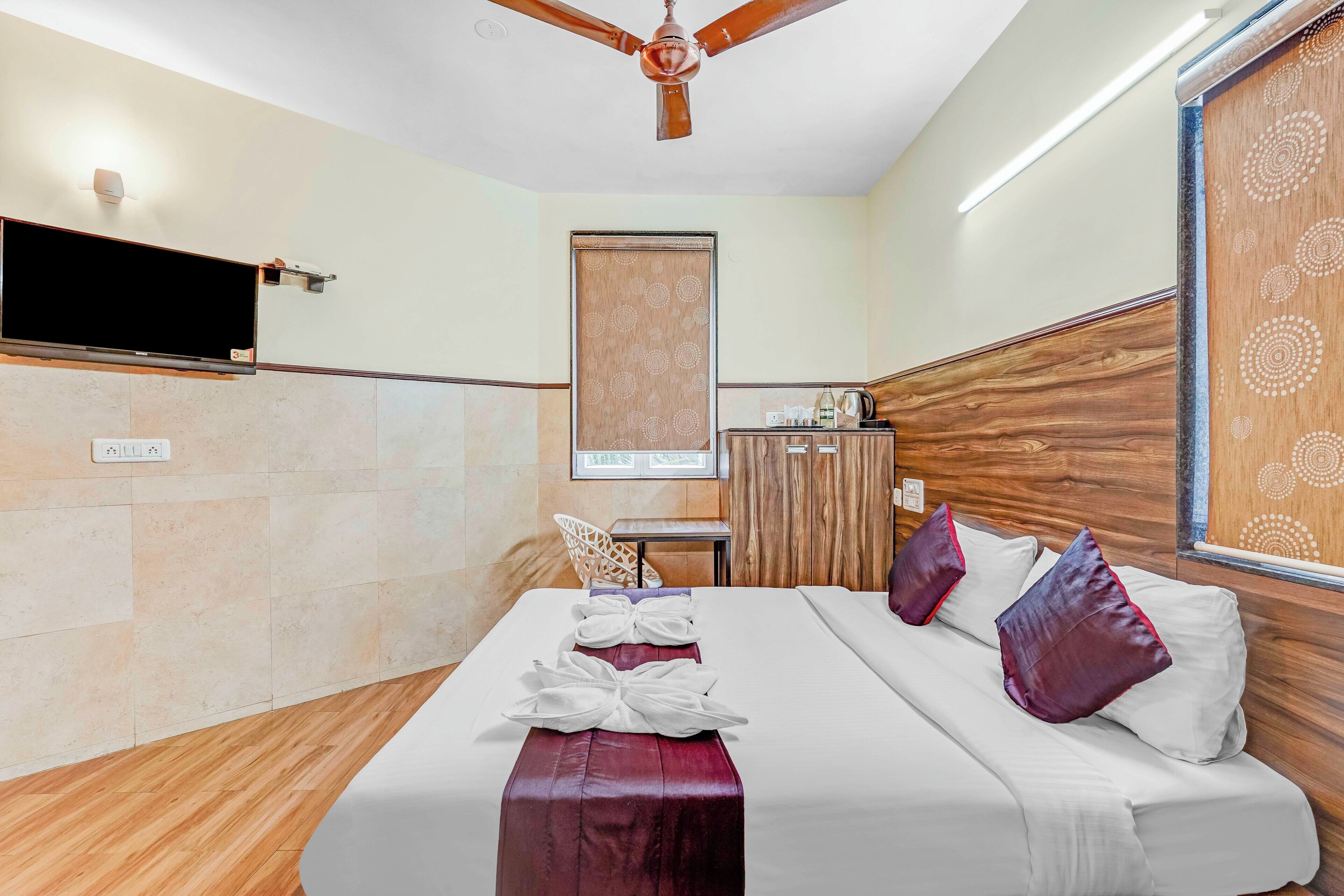 Ein Hotelzimmer mit einem Bett, einem Deckenventilator, einem Flachbildfernseher, einem Holzschrank und einem Fenster mit einem gemusterten Vorhang.